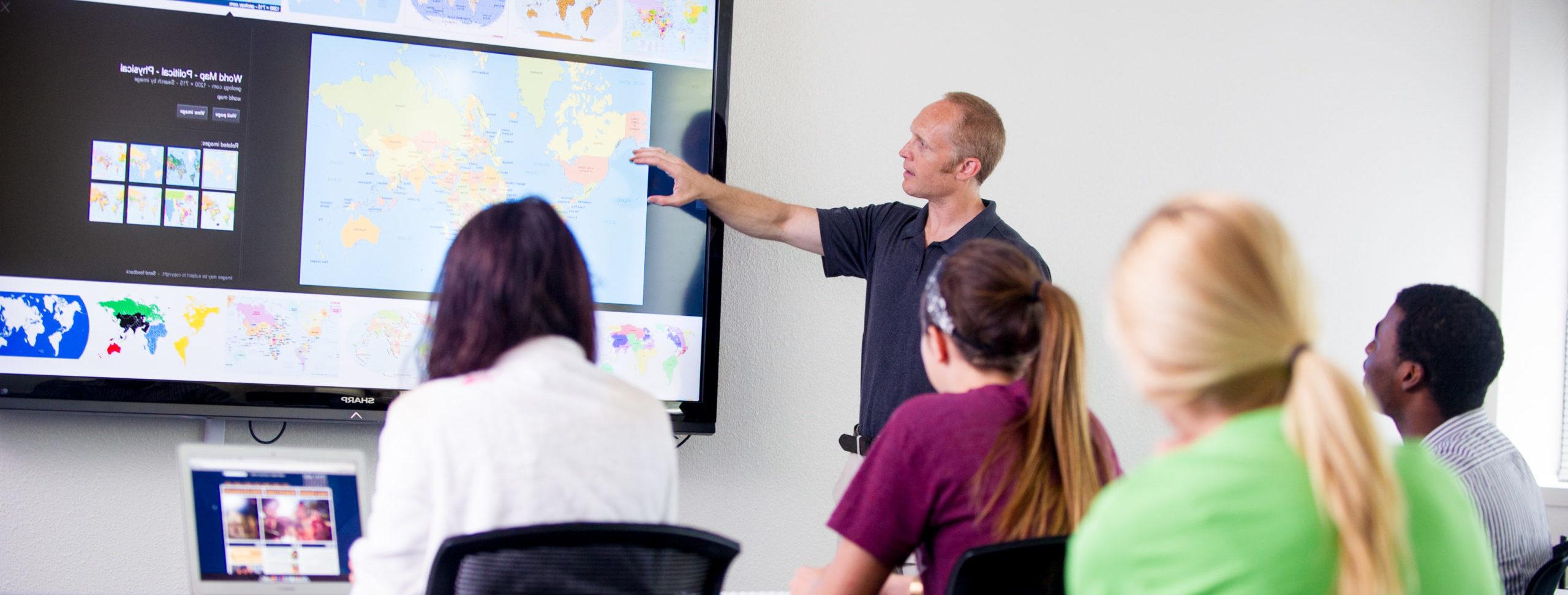 一名教师指着显示世界地图的投影仪屏幕教学生.