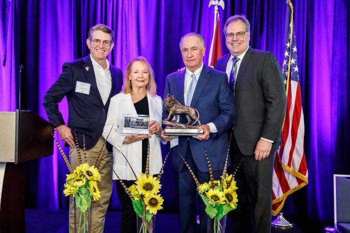 From left to right, Dr. Rudin, Senator Ted Lyon, Donna Lyon and Dr. Kelly Reyna stand on stage in front of a purple curtain and the U.S. and Texas flags. The Lyons are holding a lion statue. In front of the people, three bouquets of sunflowers sit on the floor.